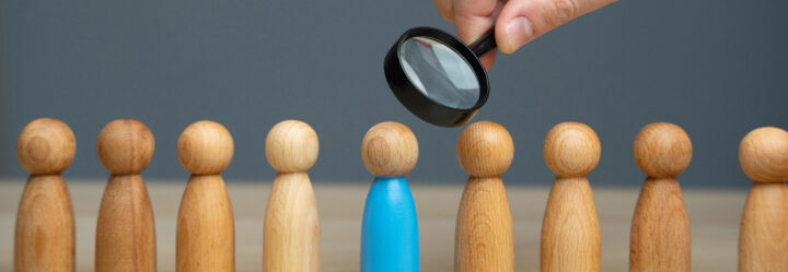 Development image concept has a man's hand holding a spyglass over a row of wooden pegs made to resemble people, and focused over one wooden peg that is painted blue.