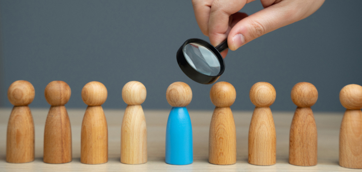 Development image concept has a man's hand holding a spyglass over a row of wooden pegs made to resemble people, and focused over one wooden peg that is painted blue.